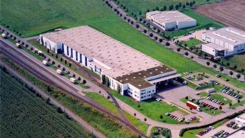 Luftaufnahme eines grossen Industriegebäudes mit Bahngleisen, LKWs und Parkplätzen.