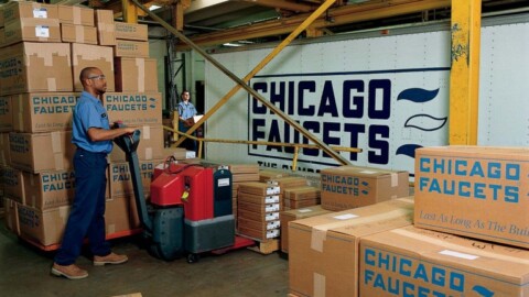 Ein Mann, der eine Hubpalette voller gestapelter Kisten der Marke "CHICAGO FAUCETS" in einem Lager bewegt.