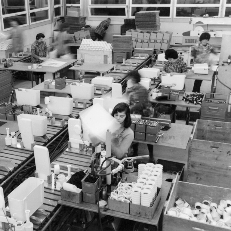 Industriehalle mit Förderbändern, an denen Mitarbeitende in einer Fabrik Spülkästen bearbeiten.