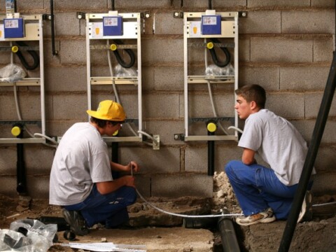 Zwei junge Handwerker, einer mit gelbem Hut, arbeiten in einem Raum an der Installation von technischen Geräten an einer Wand.