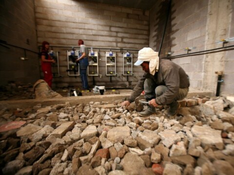 Bauarbeiter, der auf einem Boden voller Steine in einem Innenraum arbeitet, während andere Personen an Wandinstallationen arbeiten.