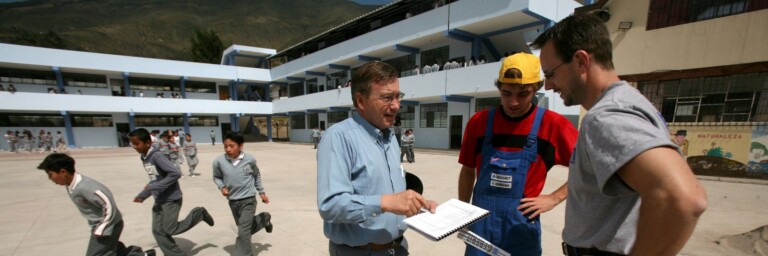 Drei Männer stehen auf einem Schulhof und diskutieren über ein Dokument, während Kinder im Hintergrund mit einem Fussball spielen.