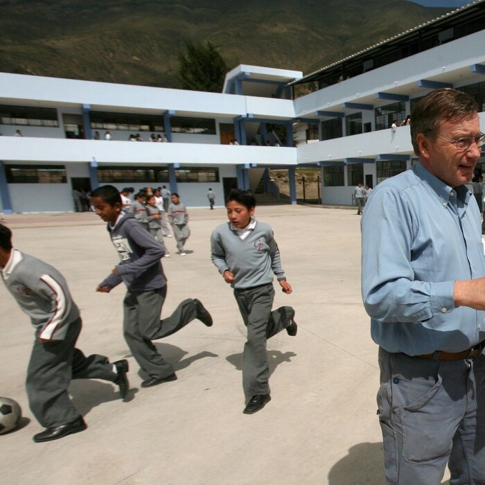 Drei Männer stehen auf einem Schulhof und diskutieren über ein Dokument, während Kinder im Hintergrund mit einem Fussball spielen.