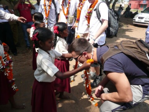 Ein Mann geht in die Hocke, während ein Kind ihm eine blumengeschmückte Kette um den Hals legt, umgeben von weiteren Kindern in traditionellen Kleidern.