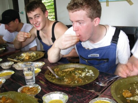 Zwei junge Männer essen gemeinsam an einem Tisch mit grossen Platten voller Essen.