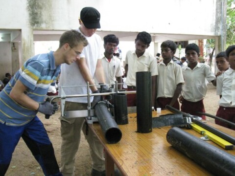 Zwei junge Männer demonstrieren eine technische Vorrichtung an einem Tisch, während mehrere Schüler in Schuluniformen aufmerksam zuschauen.