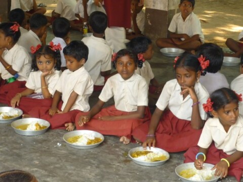 Eine Gruppe von Kindern in Schuluniform sitzt im Freien auf dem Boden und isst aus Tellern.