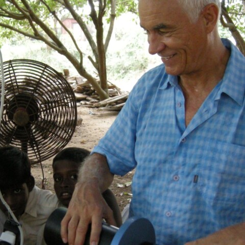 Ein älterer Mann in einem blauen Hemd steht im Freien und hält ein Rohr während eine Gruppe von Kindern ihm neugierig zuschaut. Im Hintergrund ist ein Ventilator sichtbar.