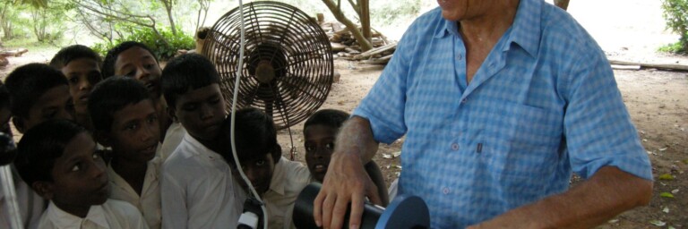 Ein älterer Mann in einem blauen Hemd steht im Freien und hält ein Rohr während eine Gruppe von Kindern ihm neugierig zuschaut. Im Hintergrund ist ein Ventilator sichtbar.