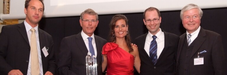 A group of five individuals posing together at an award ceremony, with one holding a trophy and another holding a large check.