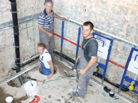 Drei Bauarbeiter stehen und sitzen in einem Arbeitsbereich mit freiliegenden Rohren und Werkzeugen.