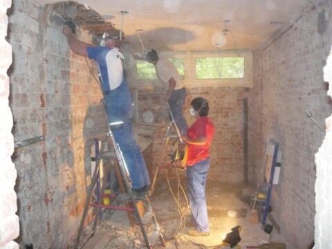 Drei Personen arbeiten in einem renovierungsbedürftigen Raum, während Staub und Schmutz in der Luft wirbeln.