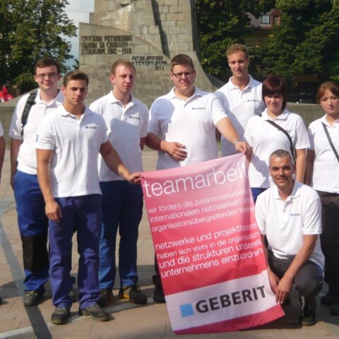 Eine Gruppe von zehn Personen in weissen T-Shirts steht lächelnd auf einem Platz, während sie ein grosses Banner halten.