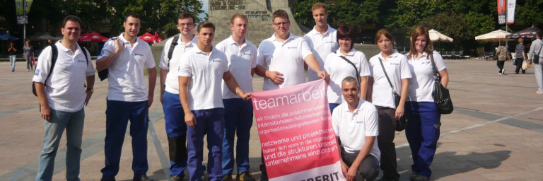Eine Gruppe von zehn Personen in weissen T-Shirts steht lächelnd auf einem Platz, während sie ein grosses Banner halten.