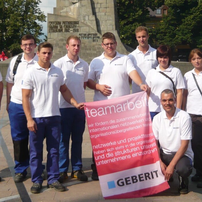 Eine Gruppe von zehn Personen in weissen T-Shirts steht lächelnd auf einem Platz, während sie ein grosses Banner halten.