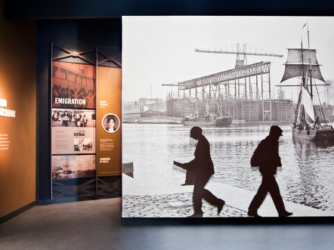 Eine Ausstellungsszene mit historischen Bildern von Passanten und einem Hafenansicht mit Schiffen.