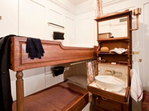 Eine holzverkleidete Kabine mit einem Etagenbett und einem integrierten Waschbecken, daneben ein Regal mit Handtüchern und Toilettenartikeln.