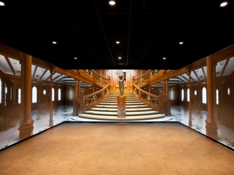 Ein Innenraum mit einem grossen, kunstvoll gestalteten Treppenhaus, das von Wandbildern umgeben ist, die die Szenen des Treppenhauses eines historischen Schiffes darstellen.