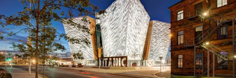 Moderne Architektur des Titanic Museums in Belfast bei Nacht, umgeben von einer Strasse und einem historischen Backsteingebäude.