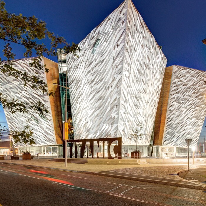Moderne Architektur des Titanic Museums in Belfast bei Nacht, umgeben von einer Strasse und einem historischen Backsteingebäude.