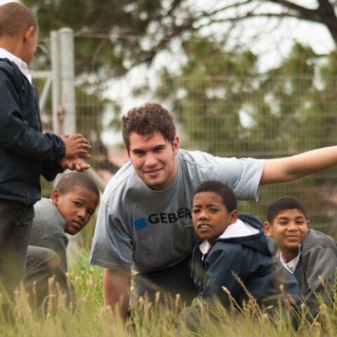 Eine Gruppe von Kindern und einem jungen Erwachsenen, die im Freien an einer Landschaftsgestaltung arbeiten, umgeben von Gras und Bäumen.
