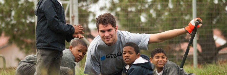 Eine Gruppe von Kindern und einem jungen Erwachsenen, die im Freien an einer Landschaftsgestaltung arbeiten, umgeben von Gras und Bäumen.