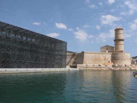 A modern black building next to a historic building by a calm harbor under a blue sky with scattered clouds.