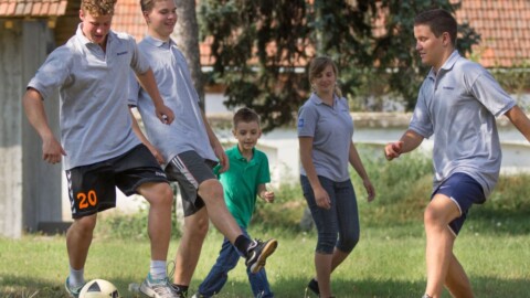 Fünf Personen spielen Fussball auf einer Wiese, umgeben von Bäumen und einem Gebäude im Hintergrund.