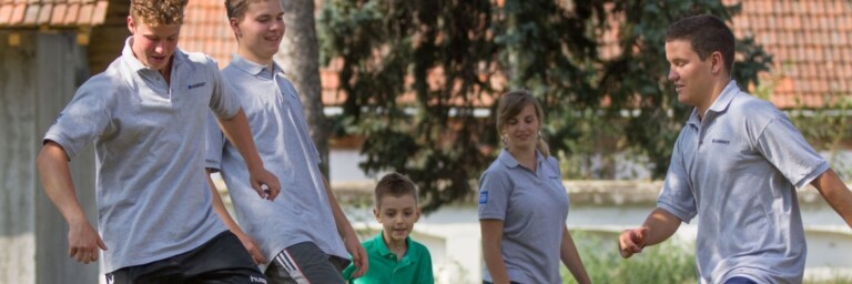 Fünf Personen spielen Fussball auf einer Wiese, umgeben von Bäumen und einem Gebäude im Hintergrund.