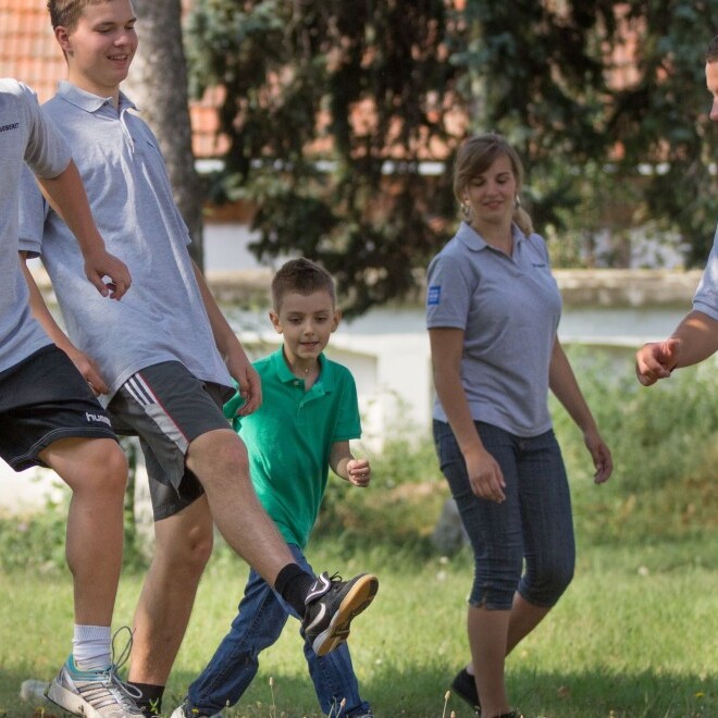 Fünf Personen spielen Fussball auf einer Wiese, umgeben von Bäumen und einem Gebäude im Hintergrund.