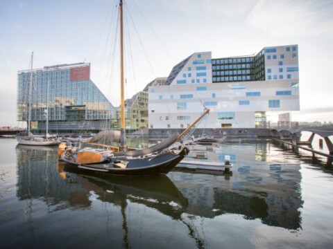 Ein historisches Segelschiff liegt im Vordergrund eines modernen Hafenbereichs mit aussergewöhnlicher Architektur und spiegelndem Wasser.