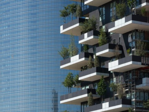 Ein modernes Wohngebäude mit mehreren Balkonen, die mit Pflanzen dekoriert sind, vor einer gläsernen Hochhausfassade.