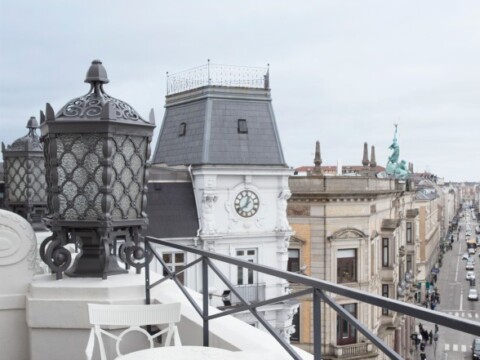 A cityscape view from a balcony featuring ornate street lamps and historic buildings, with a clock tower and sculptures visible.
