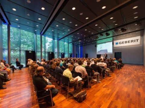 A large audience seated in a conference room with floor-to-ceiling windows and a presentation screen.