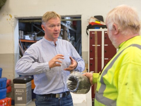 A man in a light blue shirt is gesturing while discussing with another man in a yellow reflective jacket, who is holding an object.