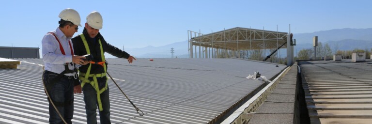 Two construction workers in safety gear inspect a rooftop.