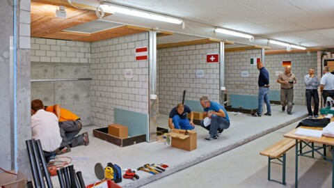 A workshop area featuring several individuals engaged in construction activities, with country flags on the walls representing Austria, Switzerland, Italy and Germany.