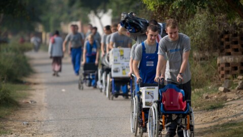 Eine Gruppe von Menschen schiebt Rollstühle auf einem unbefestigten Weg, umgeben von Grün.