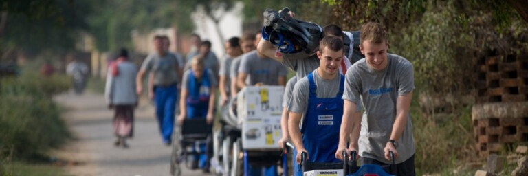 Eine Gruppe von Menschen schiebt Rollstühle auf einem unbefestigten Weg, umgeben von Grün.