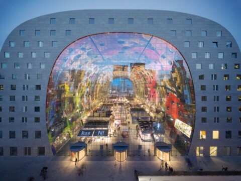 A modern building with a large, colorful archway revealing a vibrant indoor market scene, illuminated at dusk.