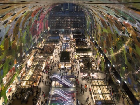 A bustling indoor market with colorful artwork on the ceiling, featuring various food stalls and shoppers navigating the aisles.