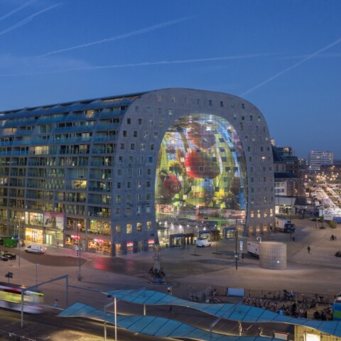 Cityscape at dusk featuring modern buildings with illuminated facades, including a large, curved structure displaying colorful artwork.