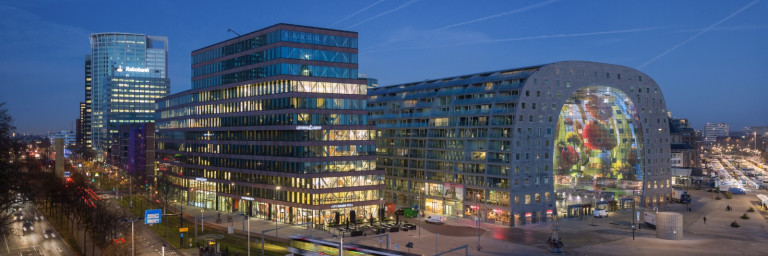 Cityscape at dusk featuring modern buildings with illuminated facades, including a large, curved structure displaying colorful artwork.