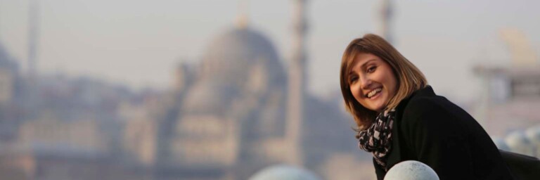 Eine lächelnde Frau in einem schwarzen Mantel lehnt an einem Geländer.