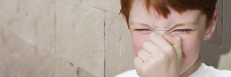 A young boy with red hair grimaces while holding his nose, standing against a textured wall.