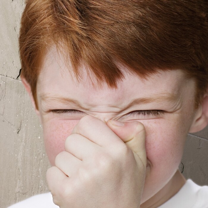 A young boy with red hair grimaces while holding his nose, standing against a textured wall.