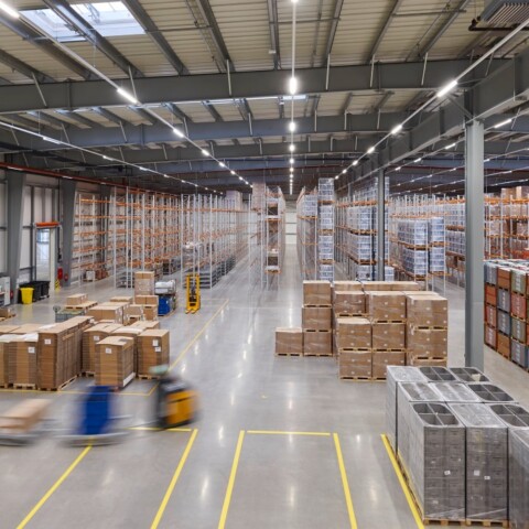 Eine grosse Lagerhalle mit hohen Regalen, auf denen Kartons gestapelt sind, und Gabelstaplern, die bei der Warenbewegung helfen.
