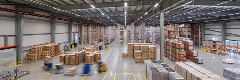 Eine grosse Lagerhalle mit hohen Regalen, auf denen Kartons gestapelt sind, und Gabelstaplern, die bei der Warenbewegung helfen.