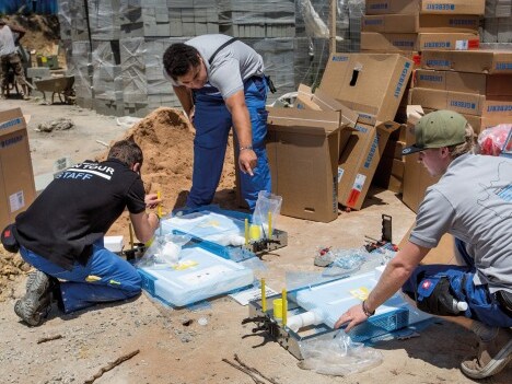 Drei Personen arbeiten auf dem Boden, umgeben von Kartons und Baustellenmaterialien, während sie mit Werkzeugen an einer Aufgabe beschäftigt sind.