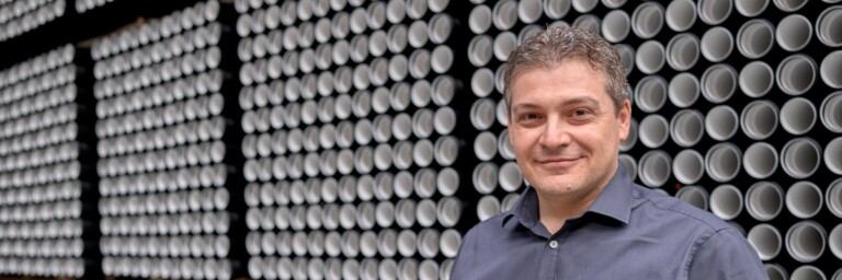 A man stands smiling in front of a wall full of pipes.